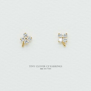 May include: Two gold-colored clover earrings, each with four clear cubic zirconia stones. The earrings are small, measuring 4 x 7 mm. The text "TINY CLOVER CZ EARRINGS" is below the earrings.