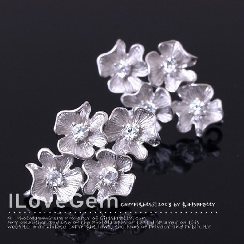May include: A silver-toned brooch or hair accessory featuring a cluster of six textured flower shapes. Each flower has five petals with a brushed metallic finish and a single clear, round-cut gemstone set in the center of each bloom.