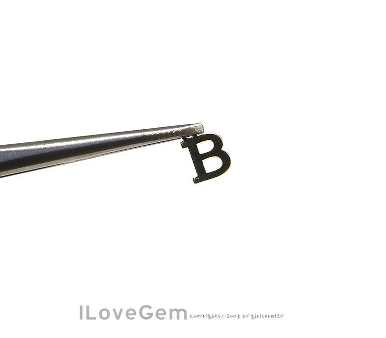 May include: A black metal letter B charm, held by tweezers.