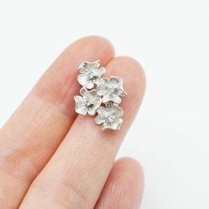 May include: A silver-toned brooch or pin featuring a cluster of four textured flower shapes, each with a small, clear, round gemstone set in the center. The metal surface has a fine, radial line pattern radiating from the center of each petal.