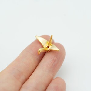 May include: A small, gold-colored metal charm shaped like an origami crane. The bird figure features smooth, polished surfaces with defined wings, a pointed tail, and a neck, with a small circular hole visible at the center of the body structure.