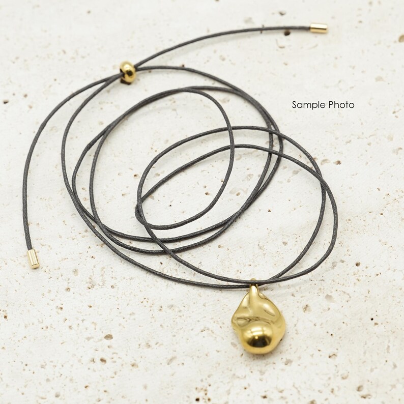 May include: A gold-colored pendant necklace with a dark gray cord. The pendant has an organic, irregular shape. The cord is adjustable with gold-colored metal accents. The words "Sample Photo" are in the background.