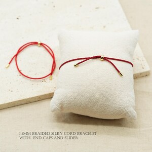 May include: Two red braided silky cord bracelets with gold-colored end caps and sliders. One bracelet is displayed on a white pillow, the other is on a light-colored surface. The text at the bottom reads: 1.5mm BRAIDED SILKY CORD BRACELET WITH END CAPS AND SLIDER.