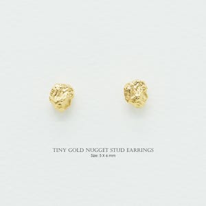 May include: A pair of tiny gold nugget stud earrings. The earrings have a textured surface and a natural, organic shape. The text below the earrings reads "TINY GOLD NUGGET STUD EARRINGS" and "Size: 5 x 6 mm".