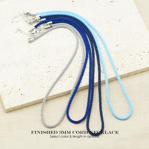 Puede incluir: Un conjunto de cuatro collares de cordón de 3mm en tonos azul claro, azul medio, azul oscuro y gris. Cada collar presenta una textura de cuerda trenzada y un cierre de metal plateado con cadena ajustable. El texto dice FINISHED 3MM CORD NECKLACE.