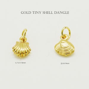 Könnte beinhalten: Zwei goldfarbene Muschel-Anhänger. Die erste Muschel ist 7,5 x 10 mm groß, die zweite 8 x 9 mm. Der Text "GOLD TINY SHELL DANGLE" steht oben im Bild. Beide Muscheln haben eine Schlaufe zum Aufhängen.