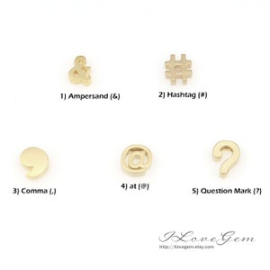 Op de afbeelding: Vijf gouden metalen bedels in de vorm van leestekens en symbolen: ampersand, hashtag, komma, apenstaartje en vraagteken.