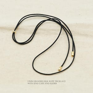 Puede incluir: Un collar de cuerda de seda trenzada negra con terminales y un deslizador dorados. El collar tiene 1,5 mm de grosor y se muestra sobre una superficie de color neutro. El texto en la parte inferior dice: 1,5 mm braided silk rope necklace with end caps and slider.