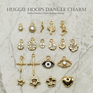 May include: A collection of 16 gold-toned dangle charms with various designs, including a cross, a star, a heart, an evil eye, a cactus, a lizard, and a peace sign. Each charm has a small loop at the top for attaching to a hoop earring.