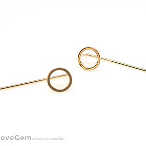 Pode incluir: Um par de brincos dourados. Cada brinco apresenta um pequeno design de círculo aberto preso a um pino reto. Os brincos estão sobre um fundo branco, destacando seu estilo simples e minimalista.