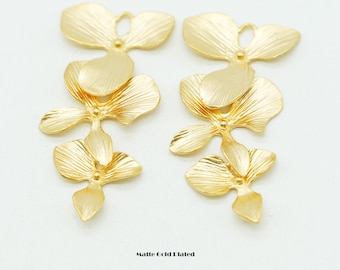 May include: Two matte gold plated metal charms, each shaped like a cascading orchid flower with three distinct petals. The surface of the gold metal features fine, linear etched textures. Each charm includes a small circular loop at the top.