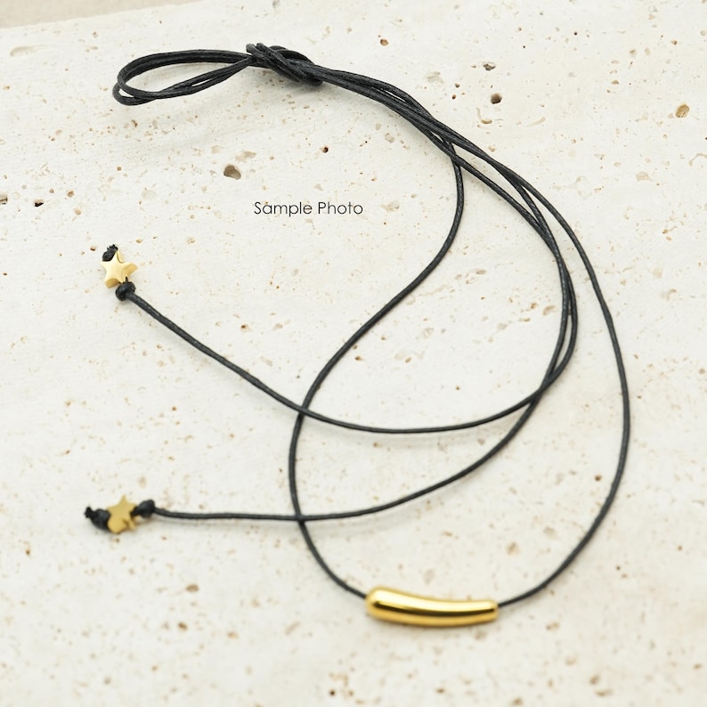 May include: Black cord necklace with a gold-colored cylindrical bead and two small star-shaped beads. The text "Sample Photo" is printed on the background.