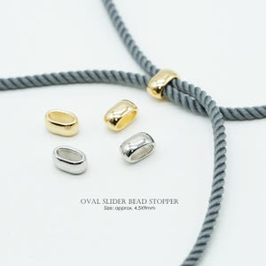 May include: Oval slider bead stoppers in gold and silver tones, featuring a smooth, rounded design. The text reads OVAL SLIDER BEAD STOPPER Size: approx. 4.5x9mm. One gold stopper is threaded onto a grey twisted cord, with three loose stoppers nearby.