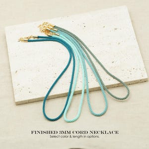 Puede incluir: Un conjunto de collares de cordón acabado de 3 mm en un degradado de tonos azules y verdes. Cada cordón cuenta con terminales de metal dorado y un cierre de mosquetón. El texto FINISHED 3MM CORD NECKLACE es visible en la imagen.