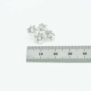 May include: A silver-toned brooch featuring four textured flower shapes, each centered with a small clear stone. The brooch is positioned above a metal ruler, showing the cluster spans approximately 1.25 inches in width.