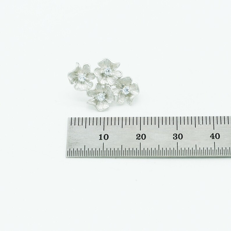 May include: A silver-toned brooch featuring four textured flower shapes, each centered with a small clear stone. The brooch is positioned above a metal ruler, showing the cluster spans approximately 1.25 inches in width.