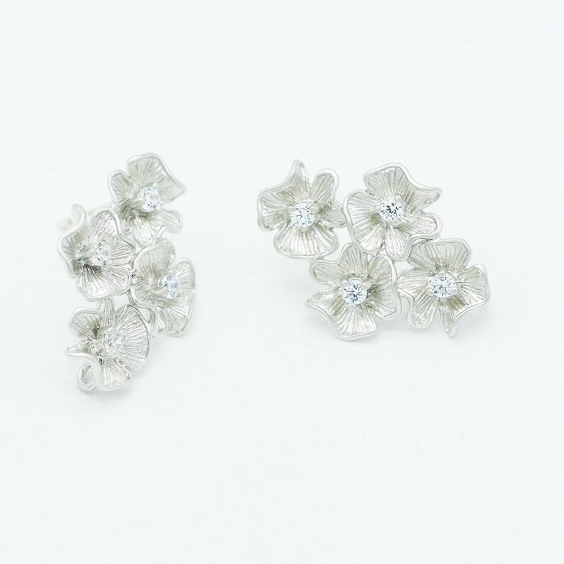 May include: A pair of silver-toned stud earrings featuring a cluster of textured, petal-like shapes arranged in a floral pattern. Each individual flower element is accented with a small, clear, round-cut gemstone set in the center of the bloom.