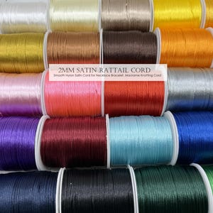 Puede incluir: Una colección de carretes de cordón cola de ratón satinado de 2 mm en varios colores, incluyendo blanco, amarillo y azul. La imagen incluye texto que dice "2MM SATIN RATTAIL CORD" y "Smooth Nylon Satin Cord for Necklace Bracelet, Macrame Knotting Cord."