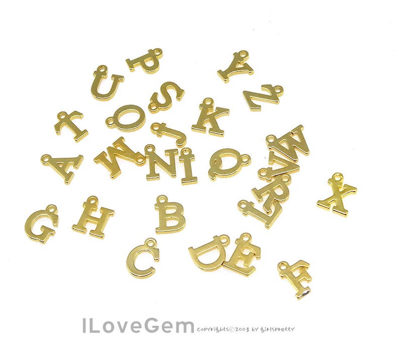 May include: A collection of gold-colored metal letter charms, each featuring a different letter of the alphabet. The charms are scattered on a white background.