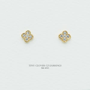 May include: A pair of gold-colored clover-shaped earrings, each with four clear, round CZ stones. The earrings are 6mm in size and are displayed on a white background. The text "TINY CLOVER CZ EARRINGS" and "Size: 6mm" are below the earrings.