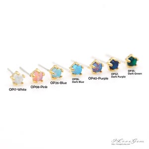 以下が含まれることがあります： 異なる色の宝石が付いた小さなスタッドピアスが8つ並んでいます。ピアスは左から右に、白、ピンク、薄い青、濃い青、紫、濃い紫、濃い緑の順に並んでいます。ピアスには、対応する色と製品コードがラベル付けされています。