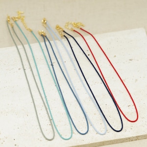 May include: A collection of six thin cord necklaces in various colors including grey, turquoise, blue, light blue, navy, and red. Each necklace features a gold-tone metal clasp and an adjustable chain link extension at the top of the cord.