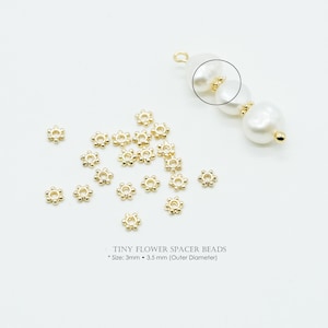Puede incluir: Pequeñas cuentas espaciadoras doradas con forma de flor esparcidas sobre una superficie blanca. Las cuentas miden 3 mm de tamaño con un diámetro exterior de 3,5 mm. La imagen también muestra un collar de perlas blancas con detalles dorados.