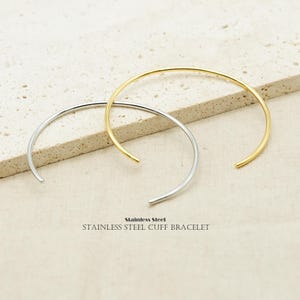 May include: Two stainless steel cuff bracelets, one silver and one gold, are displayed. The bracelets are open-ended and have a simple, minimalist design. The text "Stainless Steel Cuff Bracelet" is printed below the bracelets.