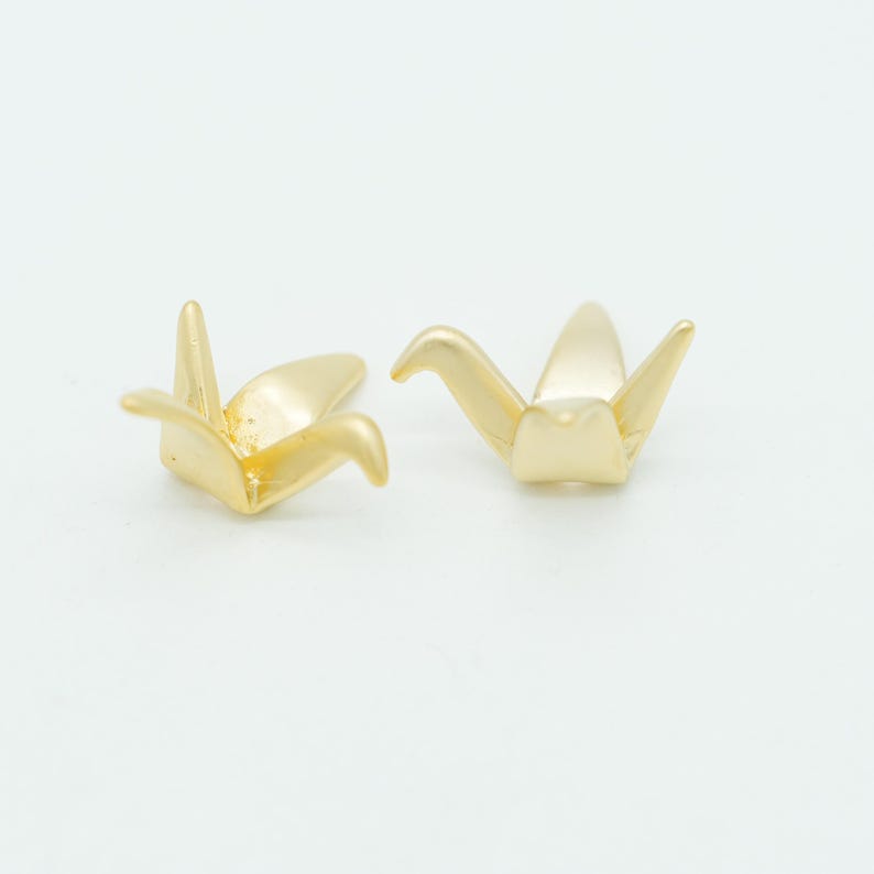 May include: Two small, gold-colored metal charms shaped like stylized origami cranes. Each piece features a smooth, reflective surface with pointed wings and a tail, designed with a hollow center. The objects are positioned side by side.
