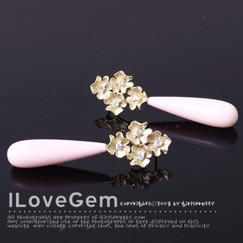 May include: A pair of gold-toned earrings featuring a cluster of three textured flowers with small clear stones at their centers, attached to a smooth, elongated, pale pink teardrop-shaped pendant. The text ILoveGem is visible at the bottom.