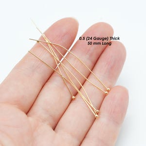 May include: A collection of gold-colored head pins, held in a hand. The pins are 0.5 (24 gauge) thick and 50 mm long. The pins have a small ball at the end of each pin. Text on the image reads: 0.5 (24 Gauge) Thick 50 mm Long.