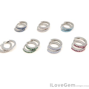 May include: A collection of small, silver hoop earrings. Each pair features a row of colorful gemstones, including blue, clear, green, light blue, and red. The earrings are displayed against a white background, showcasing their various colors and designs.
