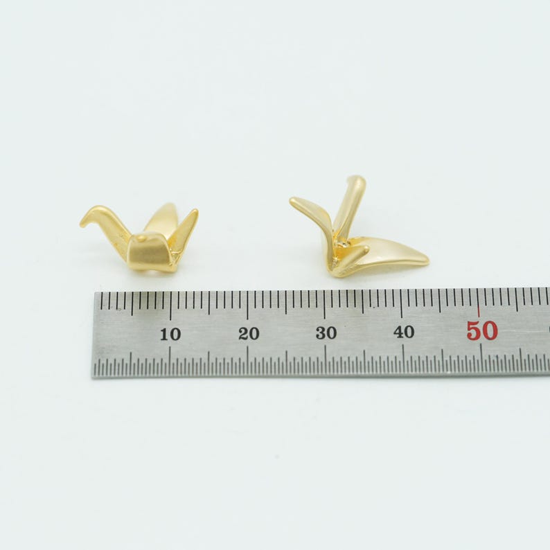 May include: Two gold-colored metal crane charms positioned above a metal ruler. The ruler shows the charms measure approximately 0.5 inches in length. The crane figures have a smooth, matte finish and are depicted in a stylized, abstract form.
