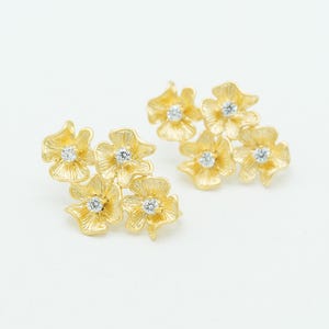 May include: A pair of gold stud earrings, each featuring a cluster of four textured flower shapes. Each individual flower petal has a brushed finish and a small, clear, round-cut gemstone set in the center, creating a bright, floral design.
