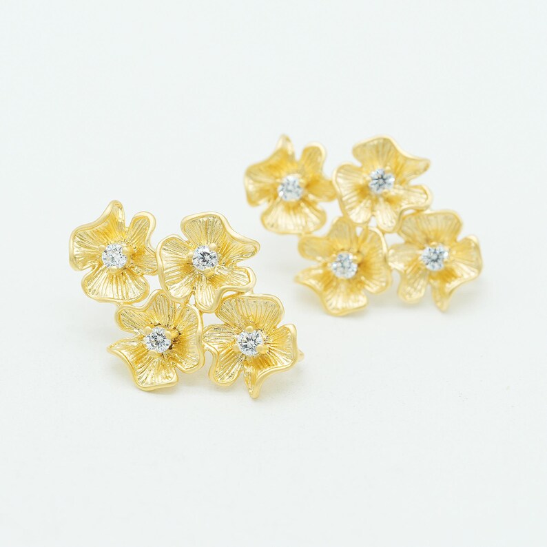 May include: A pair of gold stud earrings, each featuring a cluster of four textured flower shapes. Each individual flower petal has a brushed finish and a small, clear, round-cut gemstone set in the center, creating a bright, floral design.