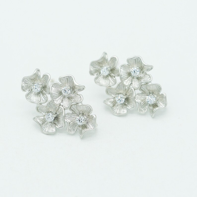 May include: A pair of silver-toned stud earrings, each featuring a cluster of four textured flower-shaped petals. Each individual flower petal has a small, clear, round-cut gemstone set in the center, creating a floral arrangement design.
