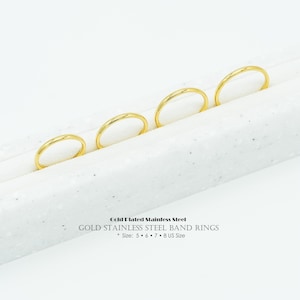 以下が含まれることがあります： 白い表面に並んだ、4つの金メッキステンレススチールバンドリング。画像には「Gold Plated Stainless Steel GOLD STAINLESS STEEL BAND RINGS * Size: 5 • 6 • 7 • 8 US Size」と書かれています。