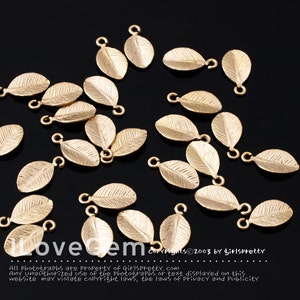 May include: A group of gold-colored metal leaf charms with a loop at the top. The leaves have a textured surface and are scattered on a black background.