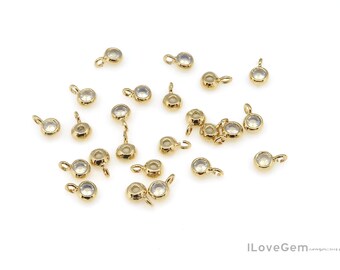 May include: A collection of small, gold-colored jewelry findings with clear, round gemstones. Each finding has a small loop for attaching to other jewelry components. The findings are scattered on a white surface.