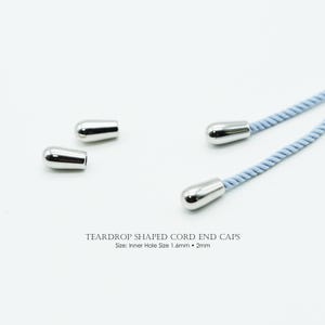 Pode incluir: Ponta de cordão prateada em forma de gota. Duas estão presas a cordões azuis claros, enquanto três estão soltas. O texto na parte inferior diz "TEARDROP SHAPED CORD END CAPS" com informações sobre o tamanho.