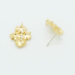 May include: A pair of gold-toned stud earrings, each featuring a cluster of three flower-shaped elements with a matte finish. The earrings include a central post backing and are designed with a textured, organic petal structure in a bright gold tone.