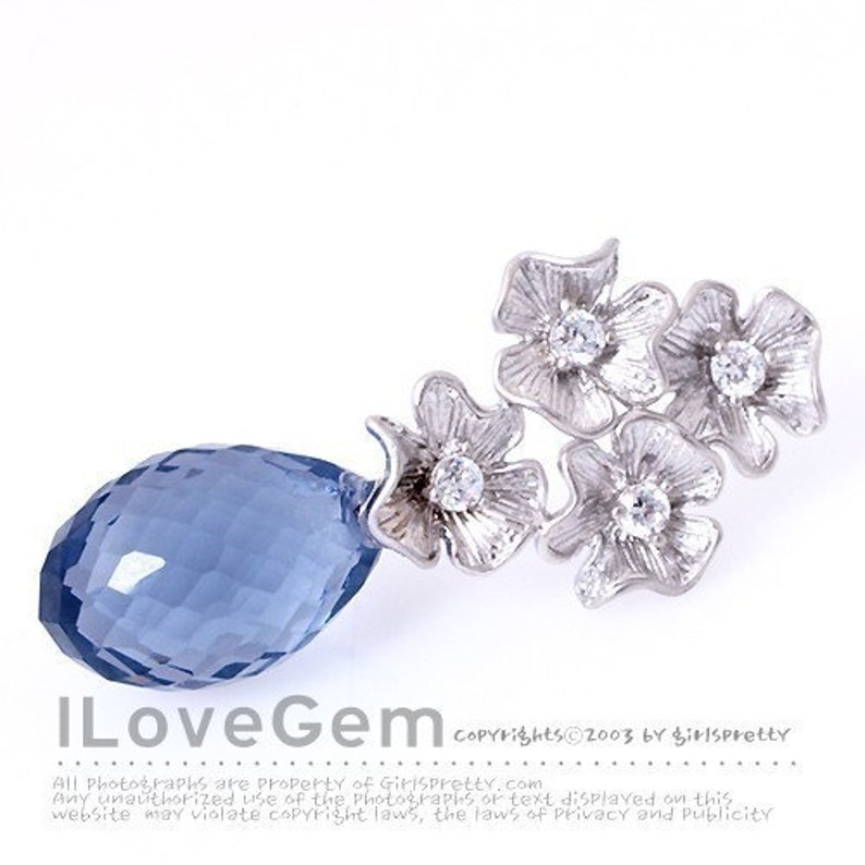 May include: A silver-toned brooch featuring a cluster of four textured metal flowers, each centered with a clear round gemstone. Attached to the floral arrangement is a large, faceted, teardrop-shaped blue crystal pendant. Text reads ILoveGem.