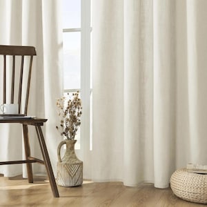 May include: Cream-colored linen curtains hang next to a wooden chair with a cup and saucer. A decorative vase with dried flowers sits near the window. The room has a natural, neutral color palette.