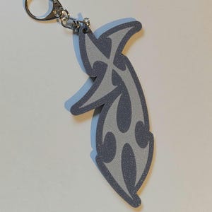 May include: A gray and silver keychain with a stylized flame design. The keychain has a silver clasp and a light blue outline. The flame design is a darker gray with a textured appearance.