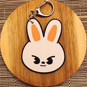 May include: A white bunny rabbit keychain with orange ears and black eyes. The bunny has a grumpy expression. The keychain is attached to a silver metal ring.