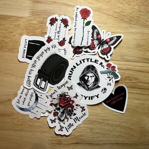 May include: A collection of stickers featuring various designs, including a skull with flowers, a belt buckle, a butterfly, a gun, a moon, and a person with a hood. The stickers are printed on white paper with black ink and have red accents.