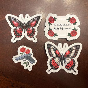 May include: Four stickers featuring butterflies and roses. One sticker has a butterfly with black and red wings. Another sticker has a butterfly with black, white, and red wings. The third sticker has a gun with red roses. The fourth sticker has red roses and the text "Emotionally attached to Zade Meadows".