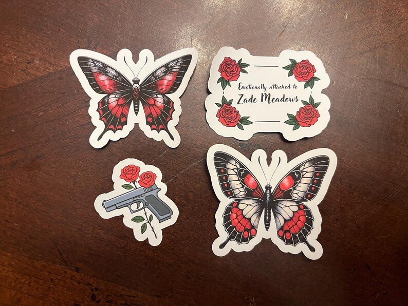 May include: Four stickers featuring butterflies and roses. One sticker has a butterfly with black and red wings. Another sticker has a butterfly with black, white, and red wings. The third sticker has a gun with red roses. The fourth sticker has red roses and the text "Emotionally attached to Zade Meadows".
