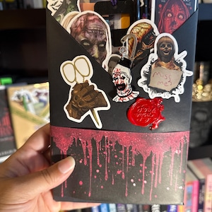 May include: A black envelope-style container decorated with horror-themed stickers and a red splatter design. Stickers include images of movie characters and a keychain. The container is held in a person's hand.