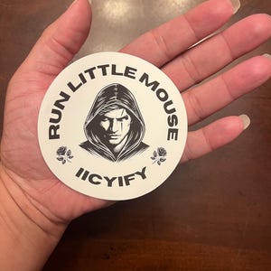 May include: A white round sticker with black text and a black and white illustration of a person wearing a hooded cloak. The text reads "Run Little Mouse Icyify" with a black rose on each side of the text.